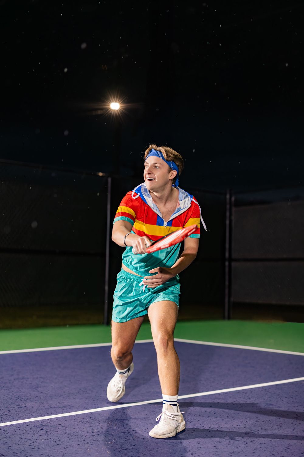 Person in colorful sportswear demonstrates athletic poses on a purple tennis court under night lights.