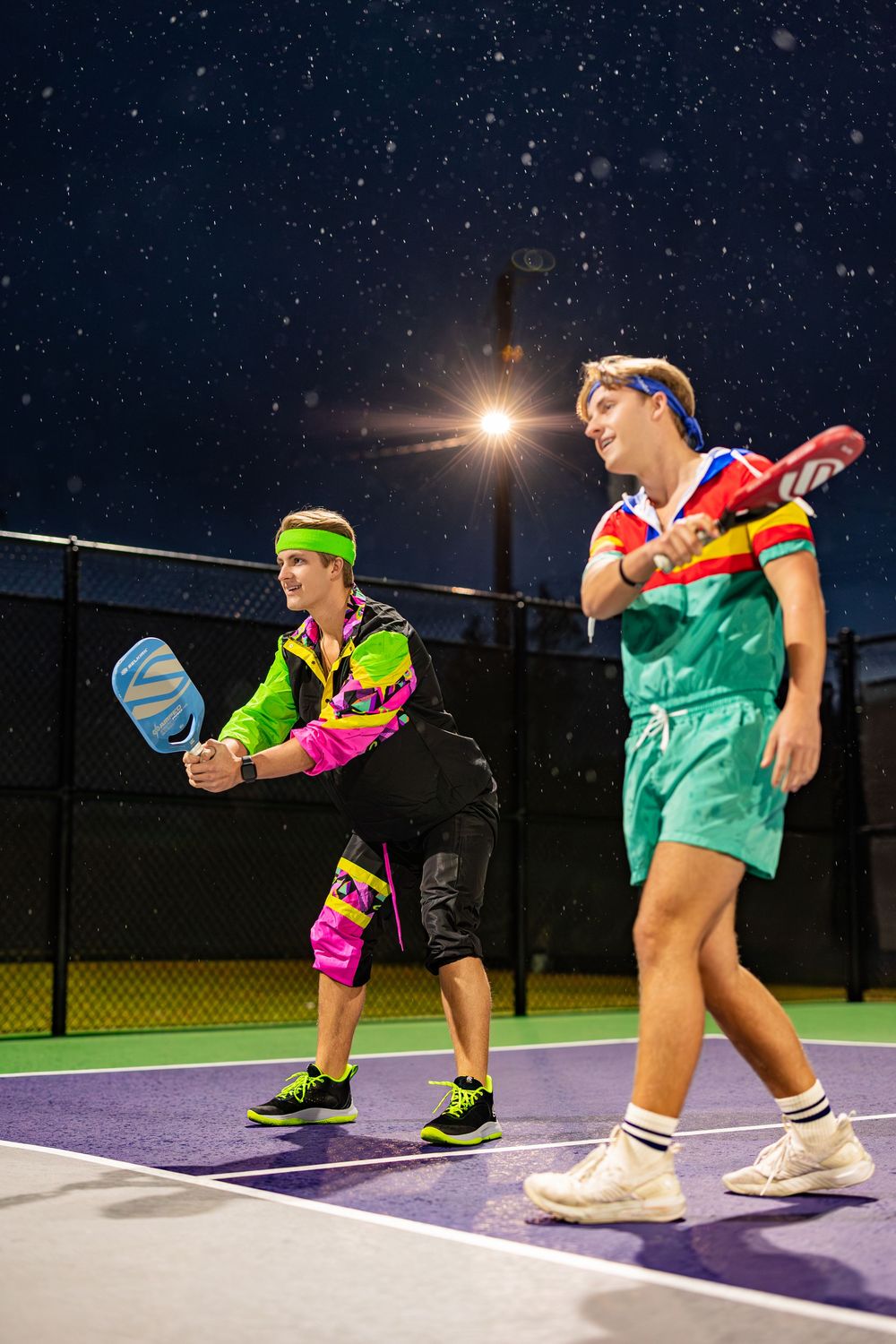 Athletes perform synchronized dance moves on a tennis court at night wearing vibrant athletic gear.