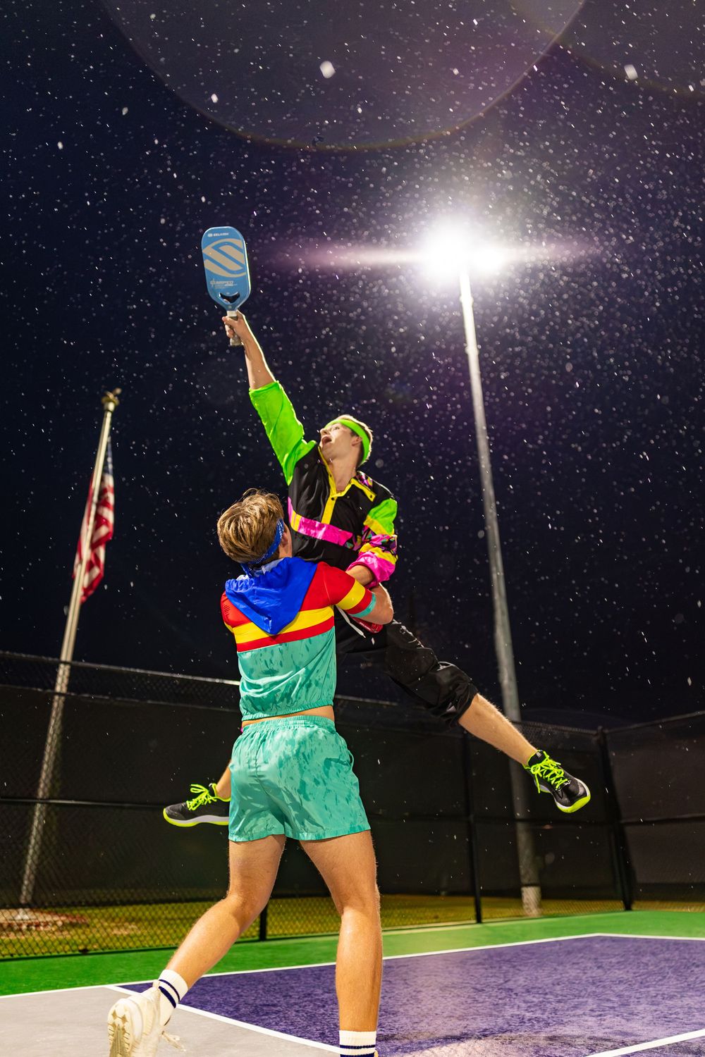 Tennis player practicing serves at night under stadium lights on an outdoor court.