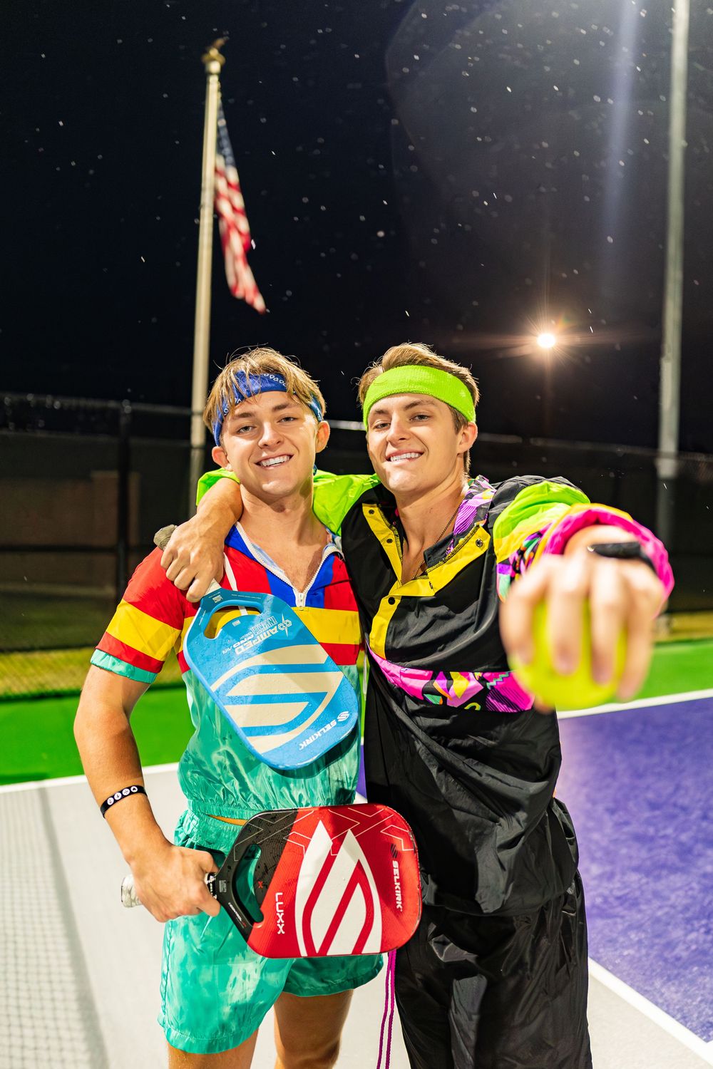 Two athletes share a joyful moment on a tennis court at night wearing colorful athletic gear.