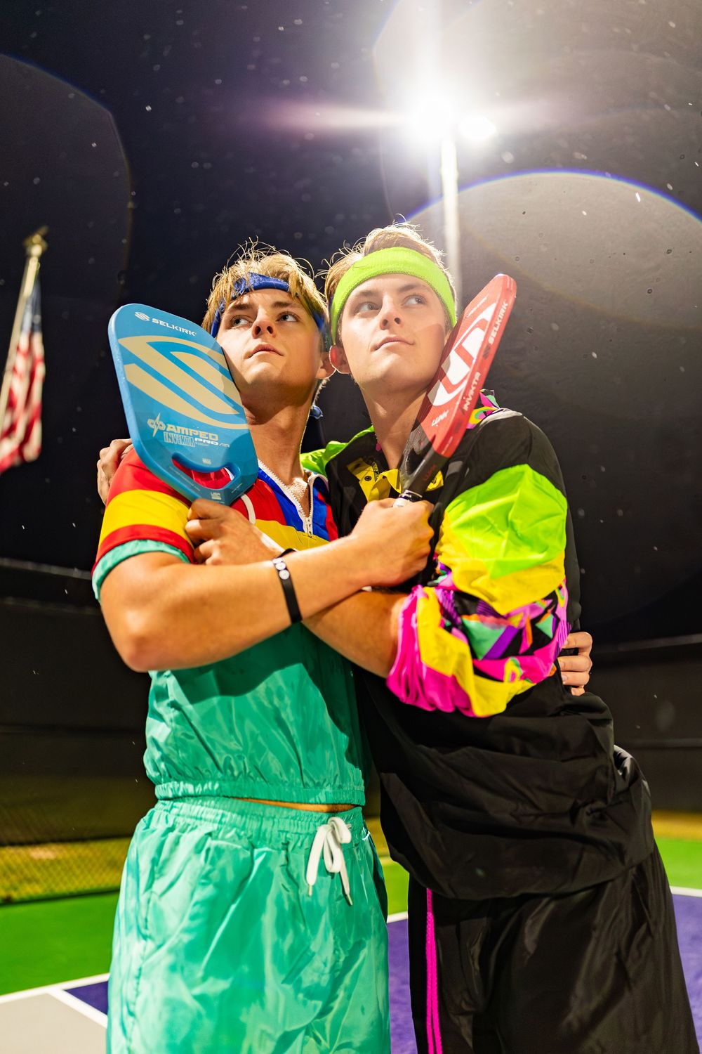 Two performers in colorful athletic wear embrace during a nighttime tennis court performance.