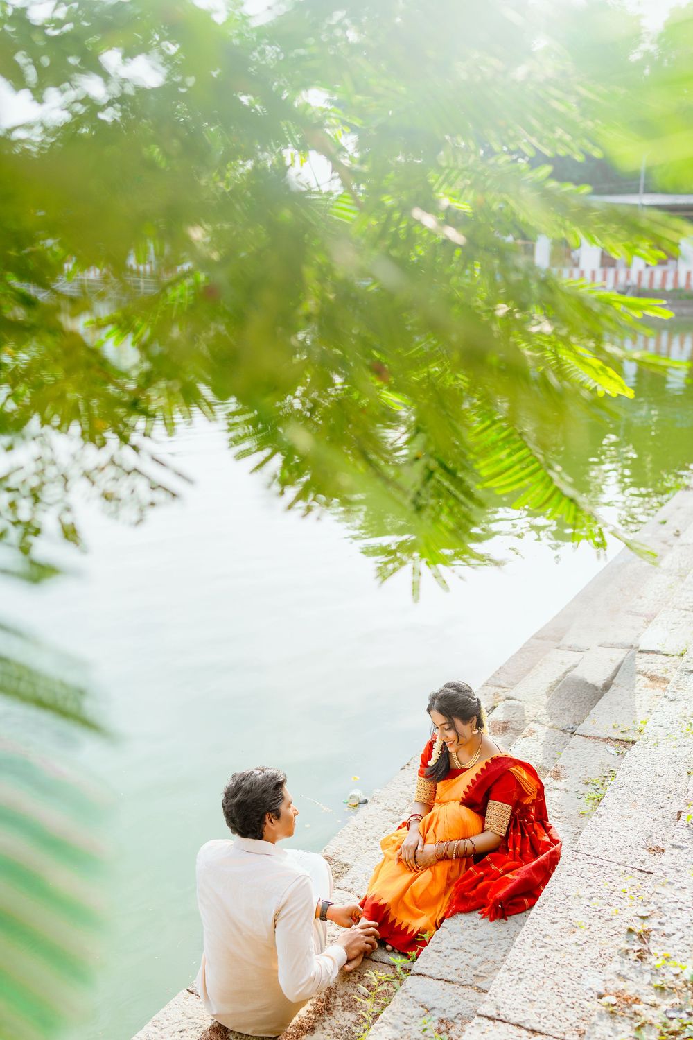 Couple in traditional Indian attire sitting by a serene pond surrounded by lush greenery.