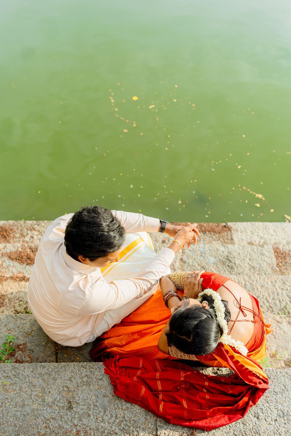 A couple in traditional Indian attire performs a ritual ceremony by a green waterfront.