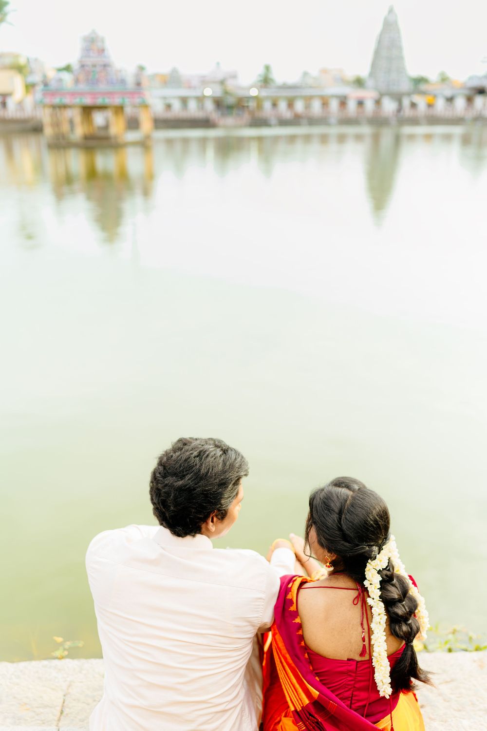 Couple in traditional dress sitting by a temple pond with buildings reflecting in the water.