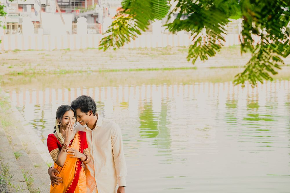 Romantic pair in traditional Indian dress embrace under green foliage near a peaceful waterfront.