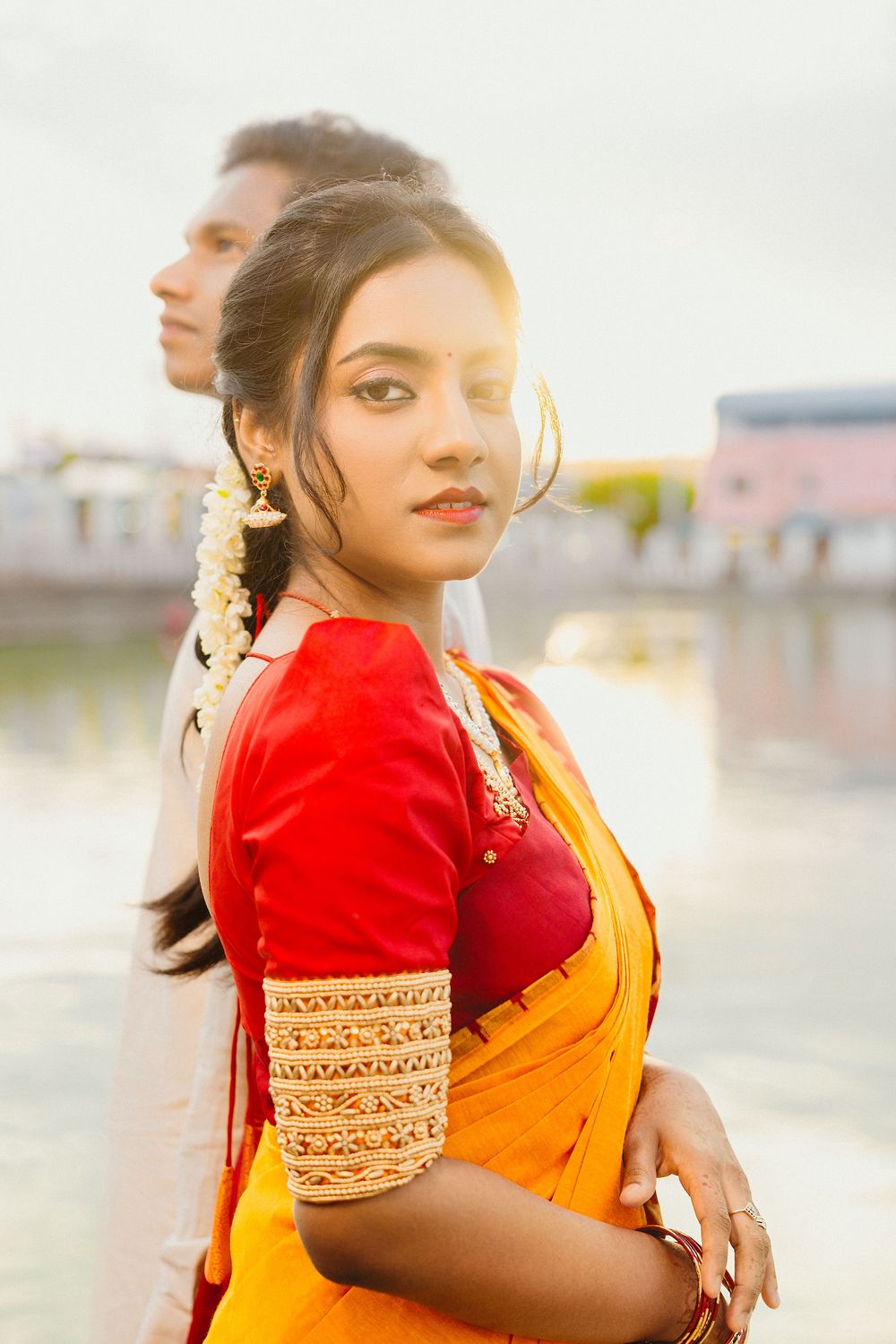 Woman in red and orange saree poses gracefully during outdoor photoshoot.