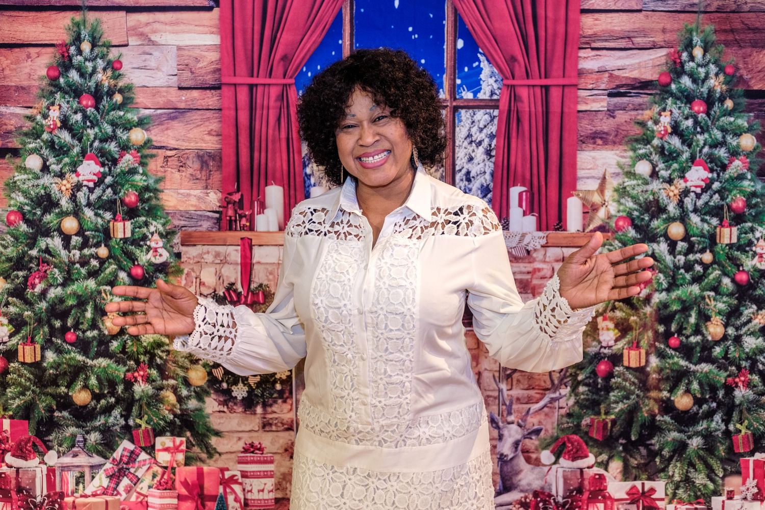 A festive holiday scene with decorated Christmas trees, red curtains, and someone in a white embroidered dress smiling.