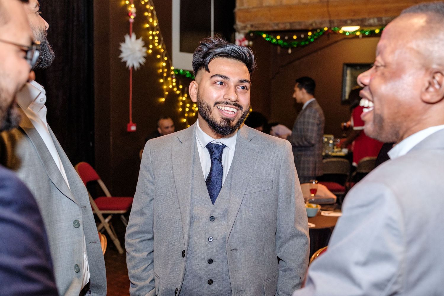 A group of people in gray suits sharing a laugh at an elegant indoor event with holiday decorations.