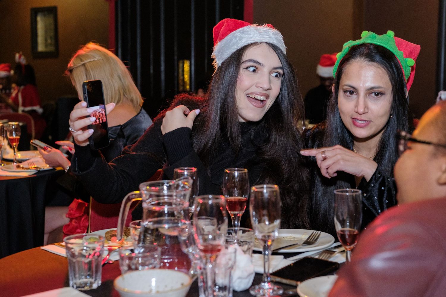 People gather around tables at a festive holiday party with drinks and Christmas decorations visible.