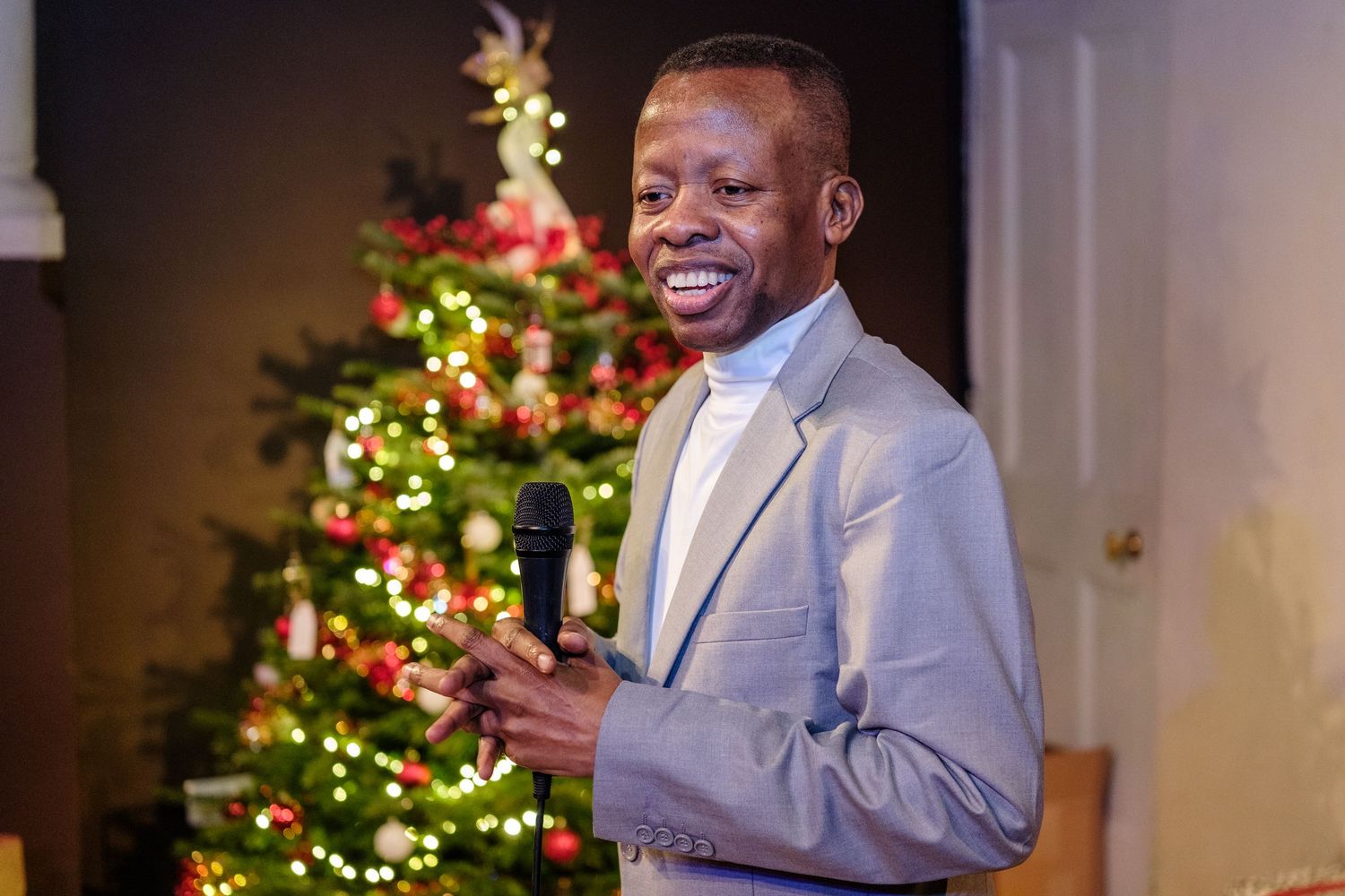 A person in a light colored suit speaks at a podium with a decorated Christmas tree in the background.