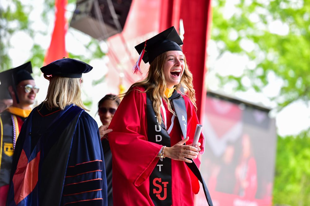 Graduates in red and black academic regalia celebrate during an outdoor commencement ceremony.