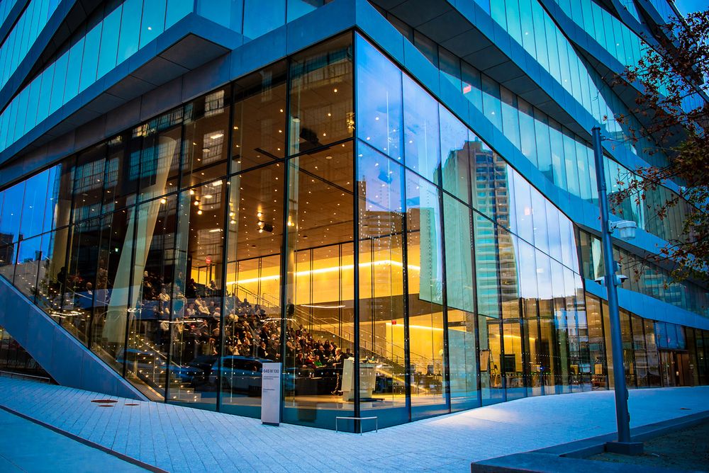 Modern glass building with angular architecture illuminated at dusk features dramatic blue and warm lighting effects.