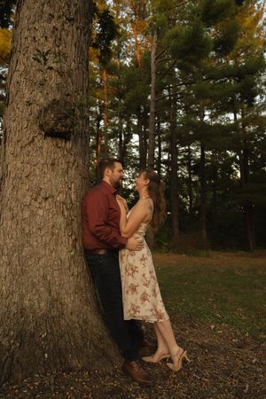 A couple poses romantically against a large tree trunk in a wooded setting during sunset.