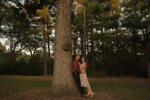 A romantic couple leans against a massive tree trunk in a forest clearing at dusk.