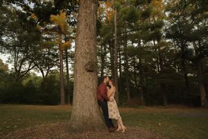 A couple shares an intimate moment against a large tree in a forest setting during golden hour.