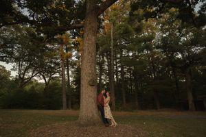 A couple stands together by a towering tree trunk in a dusky forest setting.