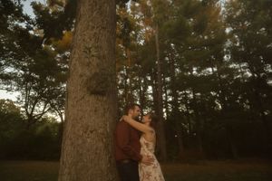 A romantic couple embraces against a large tree trunk in a wooded setting at sunset.