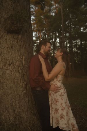 A couple shares an intimate moment leaning against a tree in a forest setting.