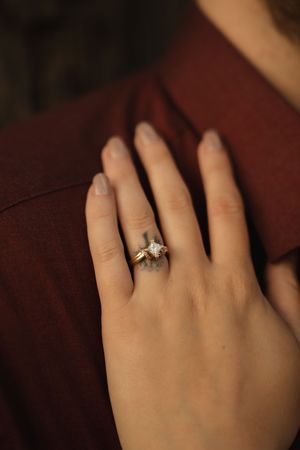 Detailed shot of a delicate engagement ring on a hand resting against dark red clothing.