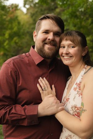 A couple poses together outdoors showing off an engagement ring.
