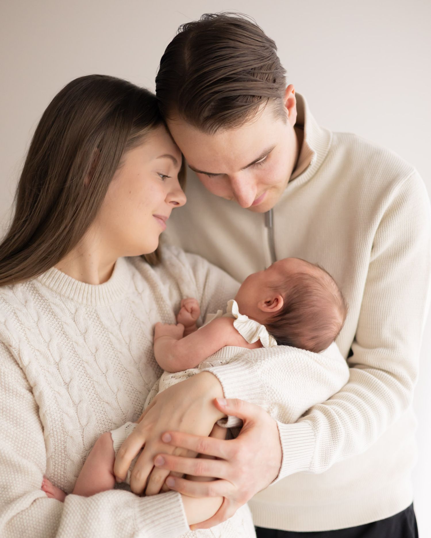 A couple in cream sweaters tenderly holds their newborn baby in an intimate family portrait.