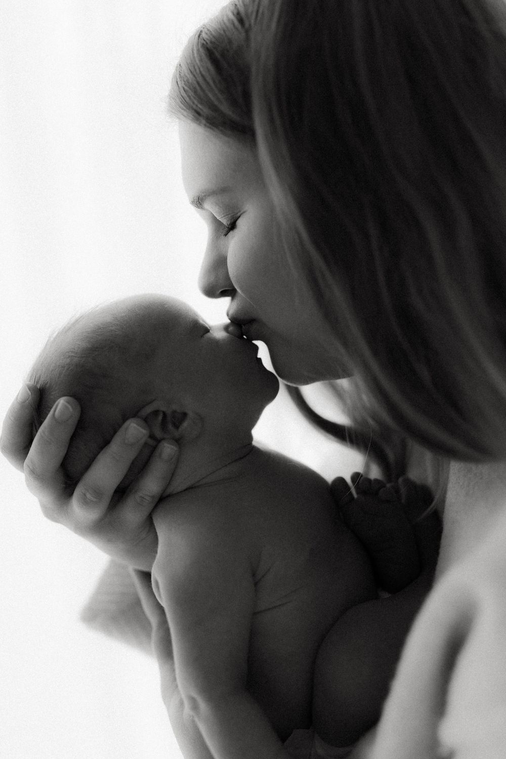 A tender black and white portrait of a mother gently kissing her newborn baby in soft lighting.