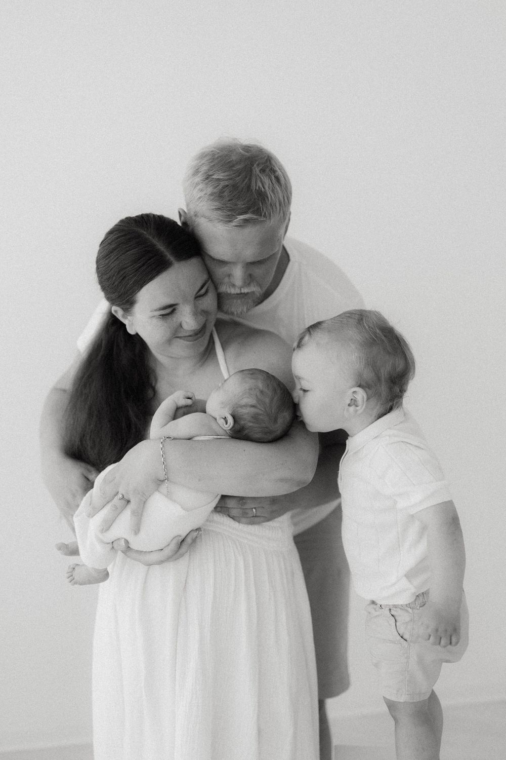 A tender black and white family portrait showing parents cradling a newborn baby while a toddler looks on.