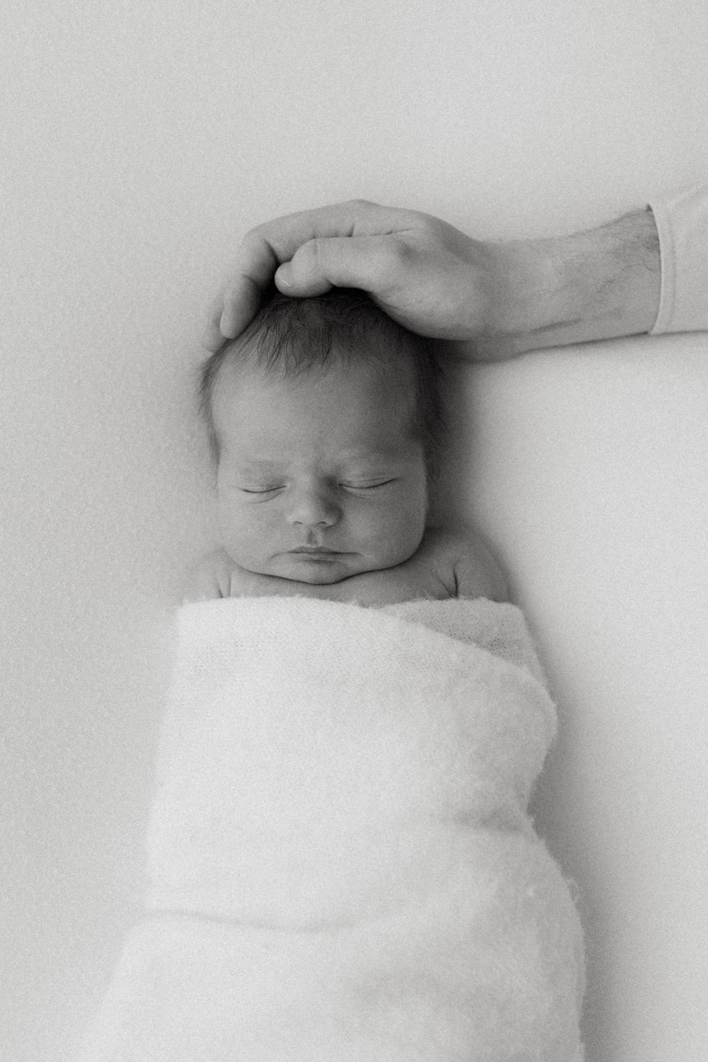 A peaceful sleeping newborn baby wrapped in white blanket with gentle hand resting on their head.