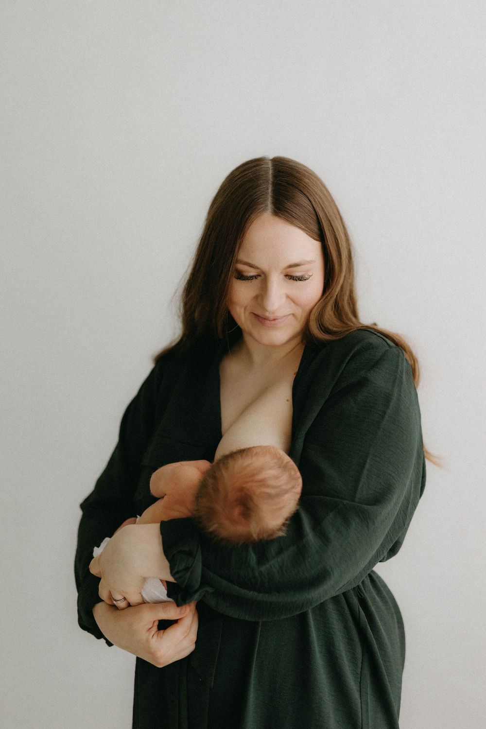 Woman in dark green robe tenderly holds and nurses her newborn baby against white wall.