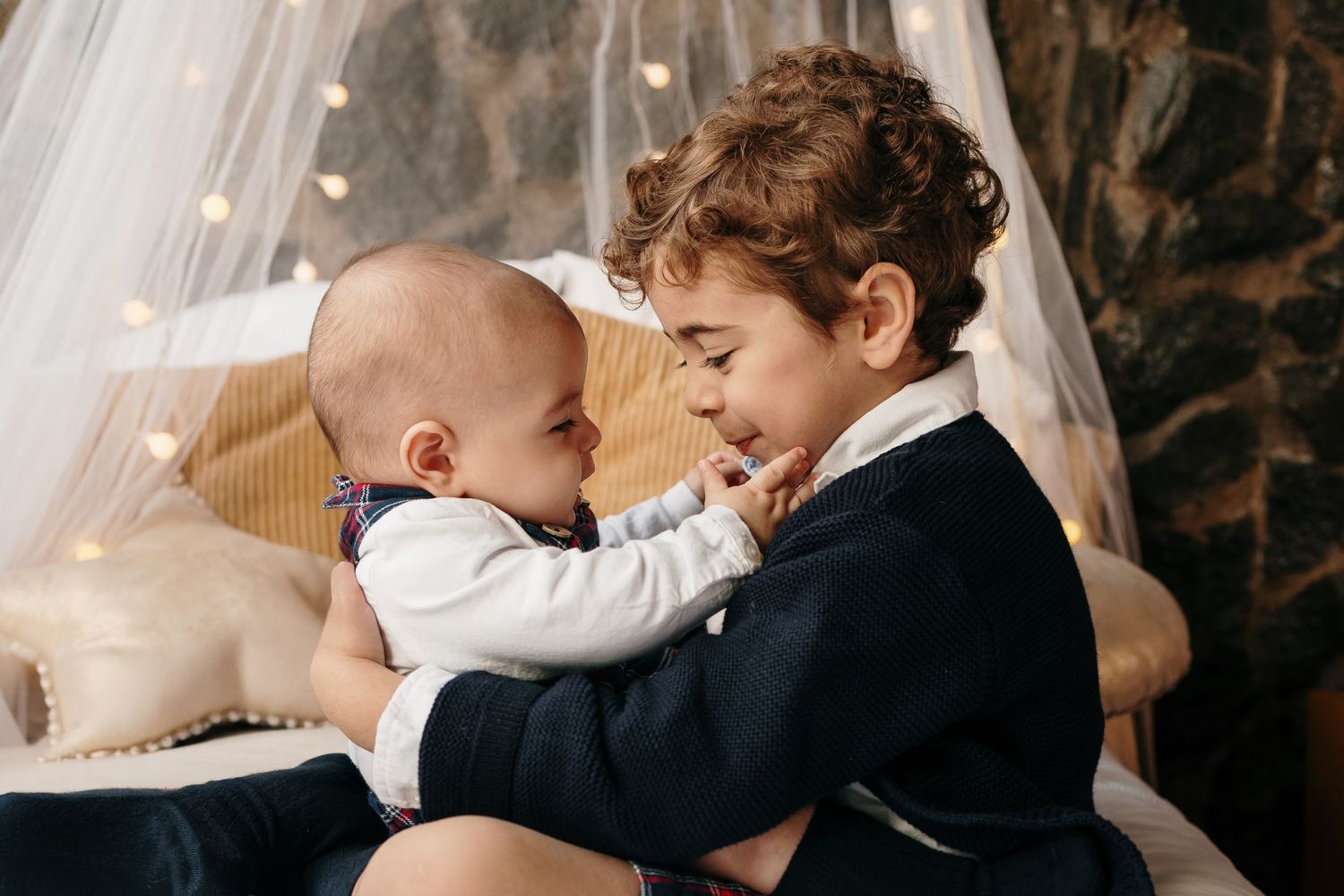 Two children sharing tender moment on plush bedding under string lights.