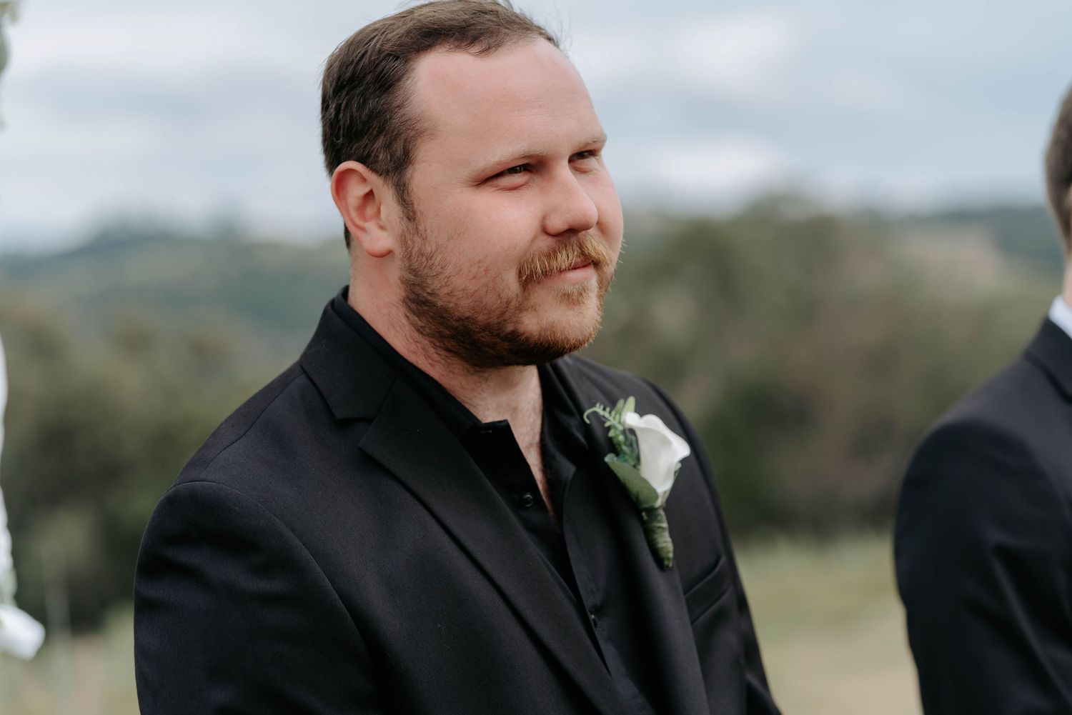 A person in a black suit smiles warmly while standing outdoors in a natural setting.