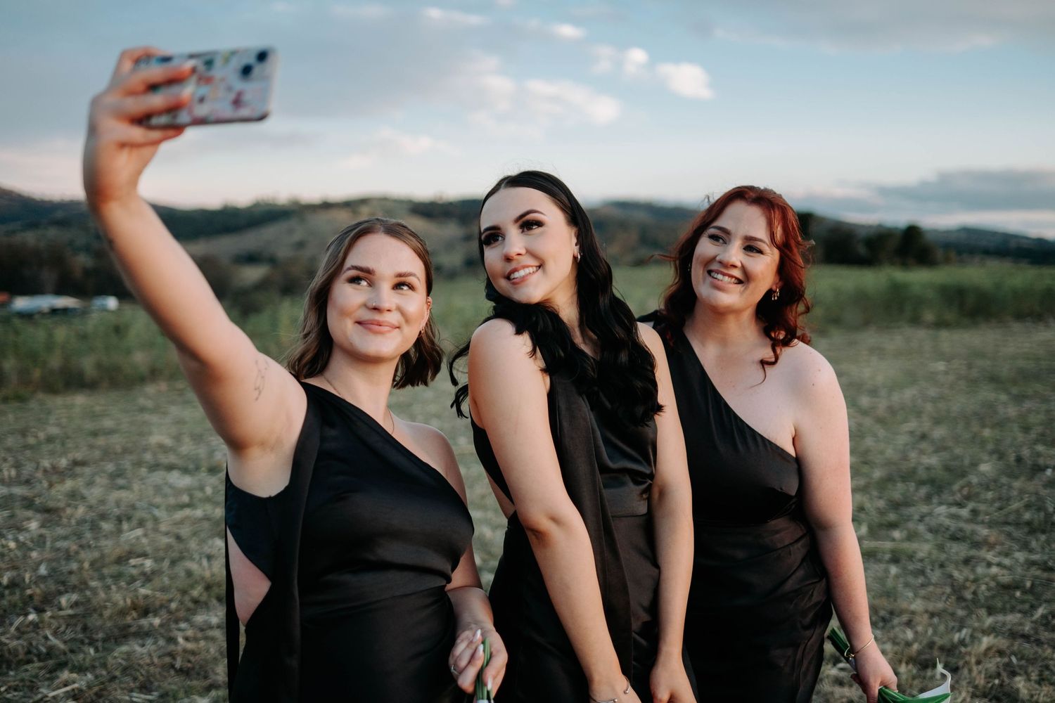 A group in black dresses poses together outdoors against a scenic mountain landscape during golden hour.