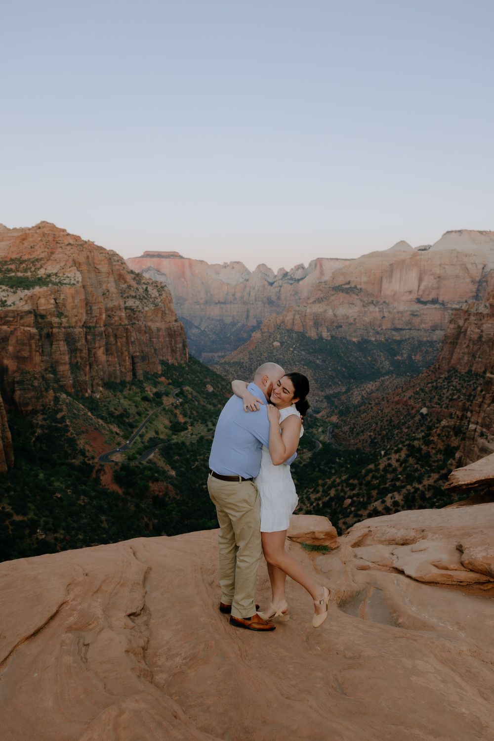 A couple embraces while standing on sandstone cliffs with a vast canyon landscape behind them.