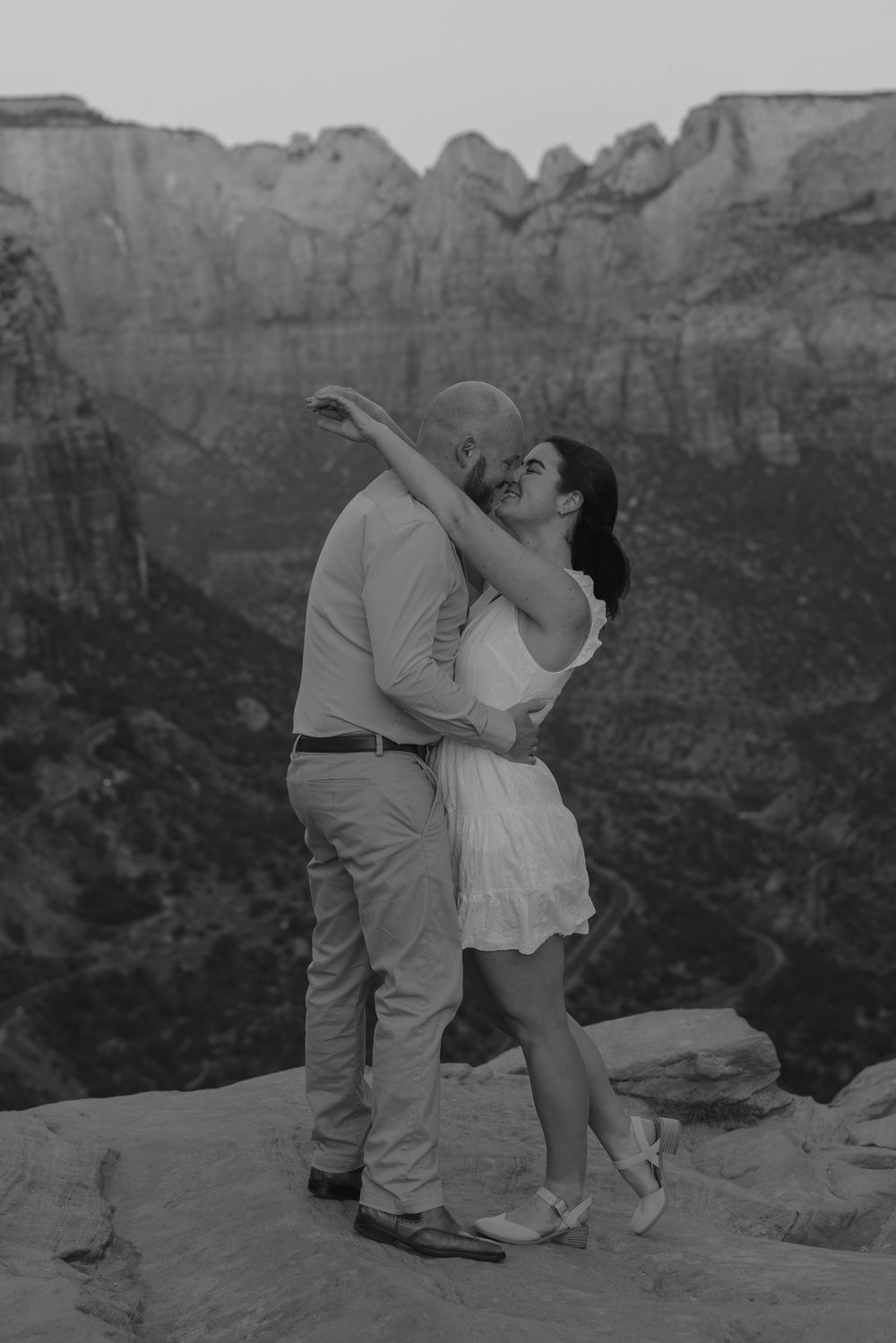 A couple shares an intimate dance pose on a desert cliff edge with towering canyon walls behind them in black and white.