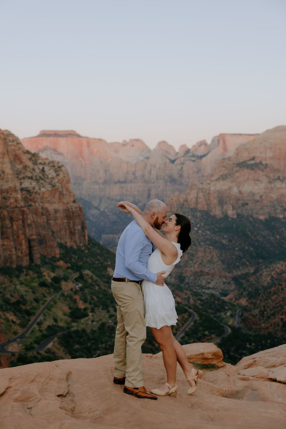A couple shares a romantic moment on red rocks overlooking the dramatic canyon landscape at sunset.