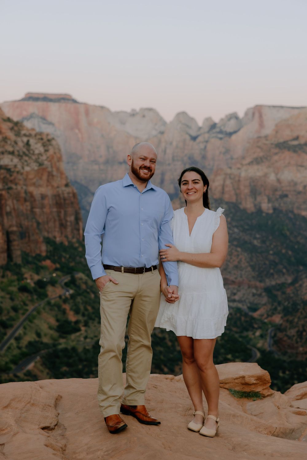 Two people pose together on a scenic overlook with dramatic red rock formations in the background.