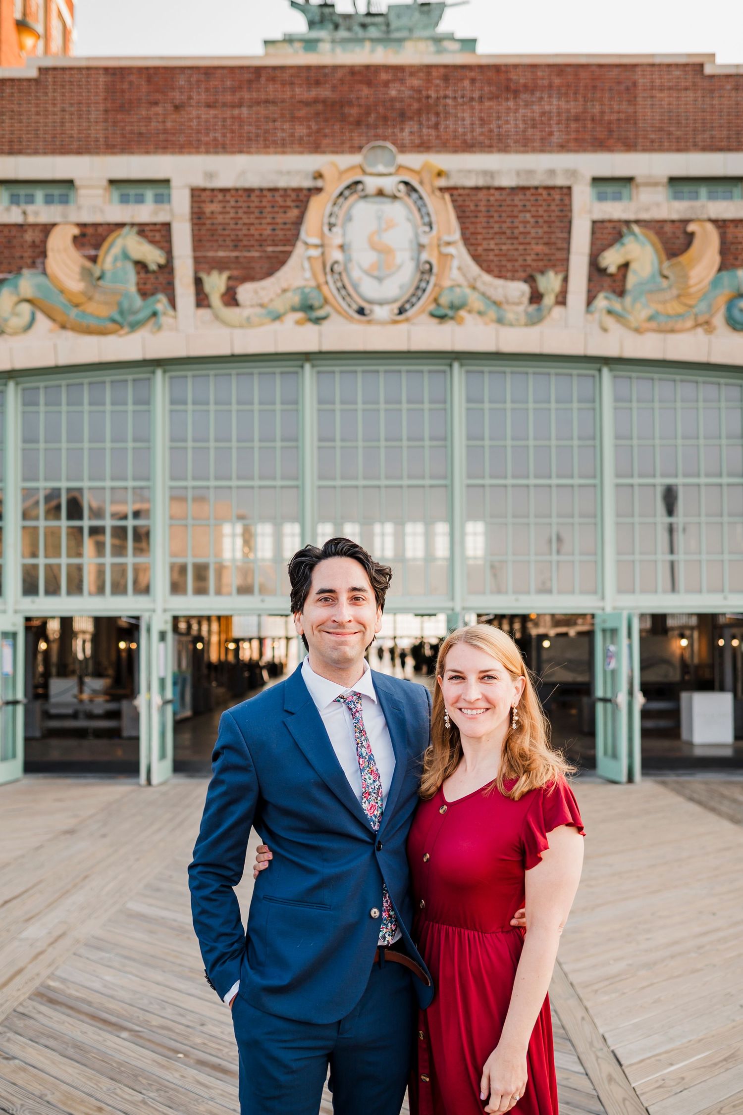 A couple poses in front of a historic brick building with ornate architectural details and large windows.