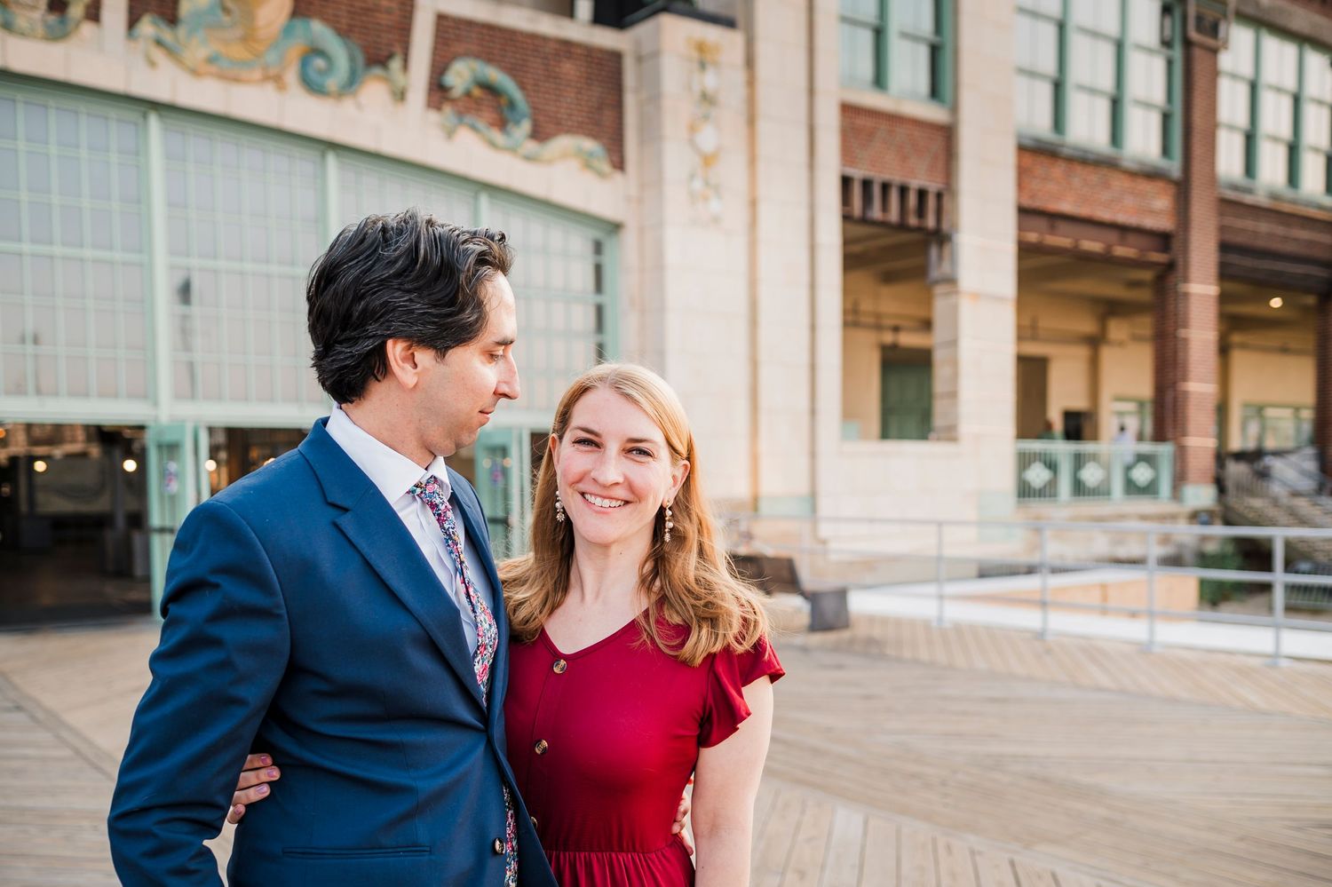 A couple in formal attire poses together outside a brick building with large windows during an engagement session.