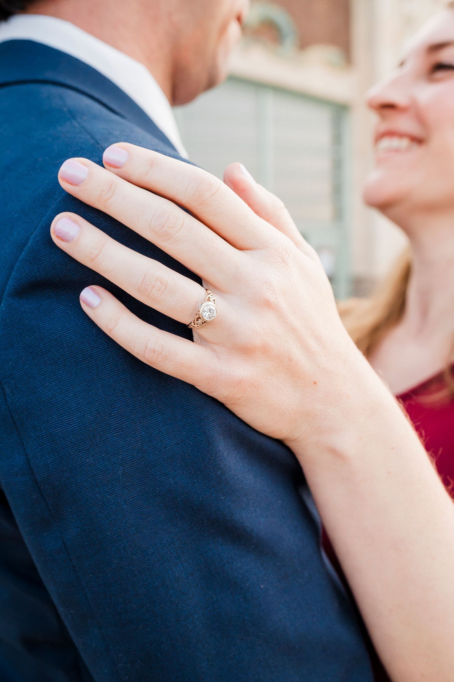 Close-up view of an engagement ring on a hand resting against a navy blue suit jacket during an intimate moment.