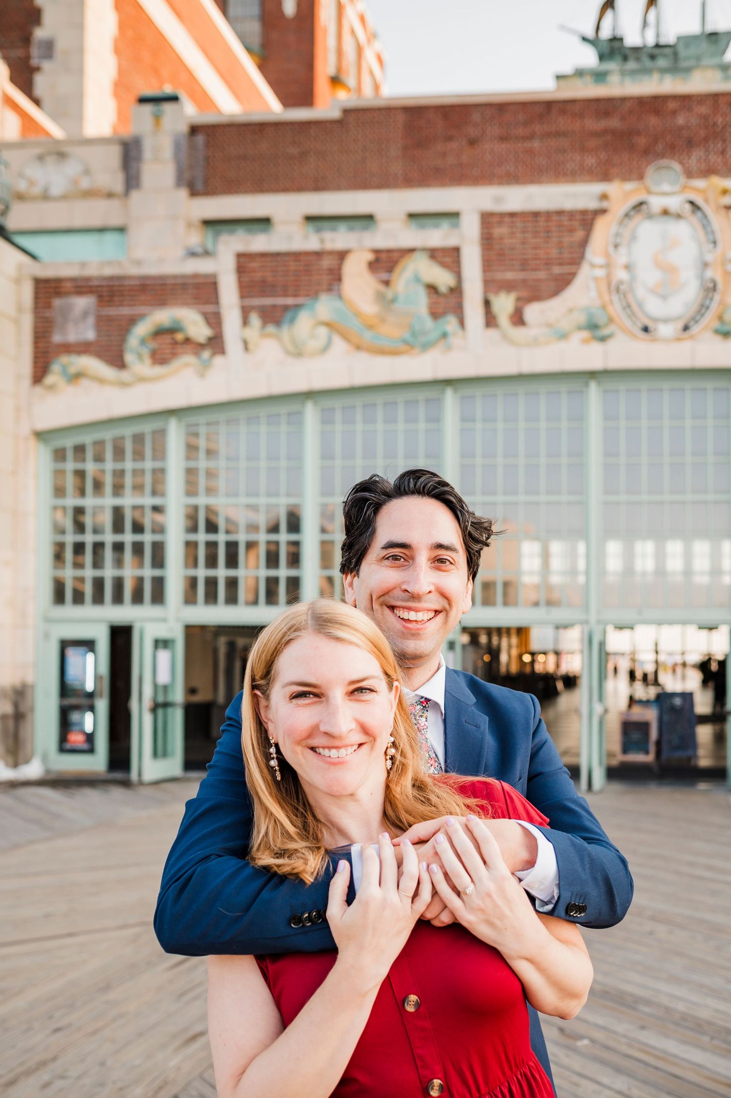A couple embraces on a boardwalk with decorative building architecture visible in the background.