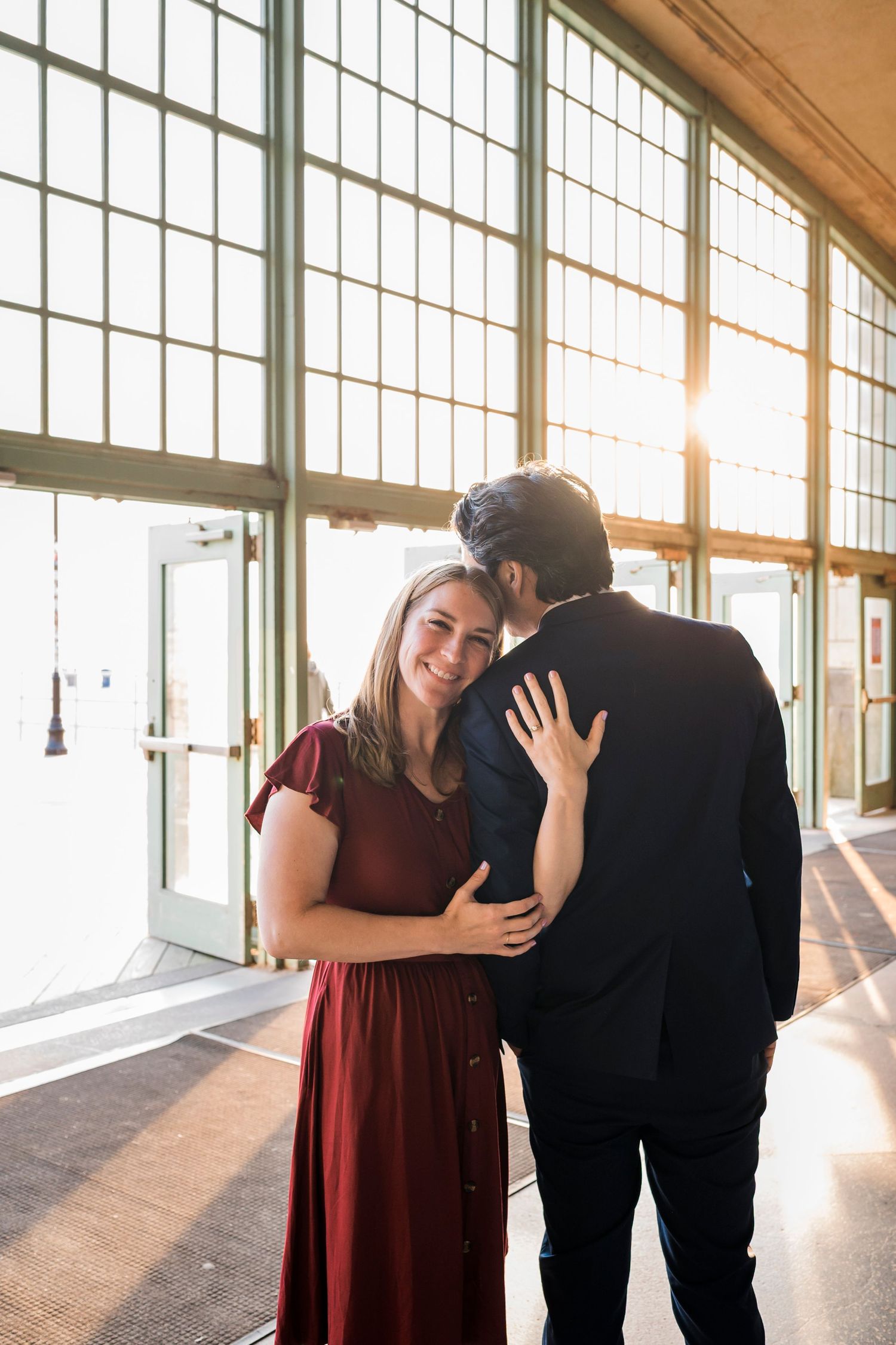 A romantic couple shares a tender moment in a sunlit hallway with large industrial windows.
