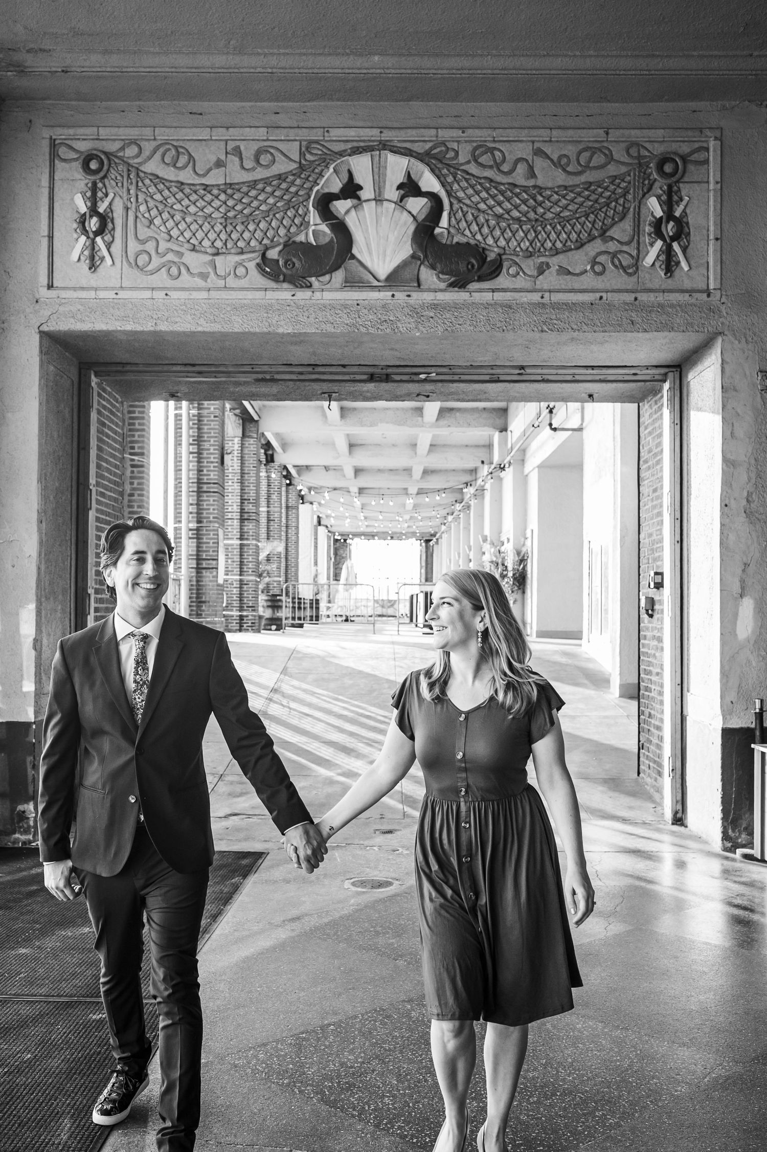 A couple walks hand in hand through an ornate colonnade hallway in formal attire.
