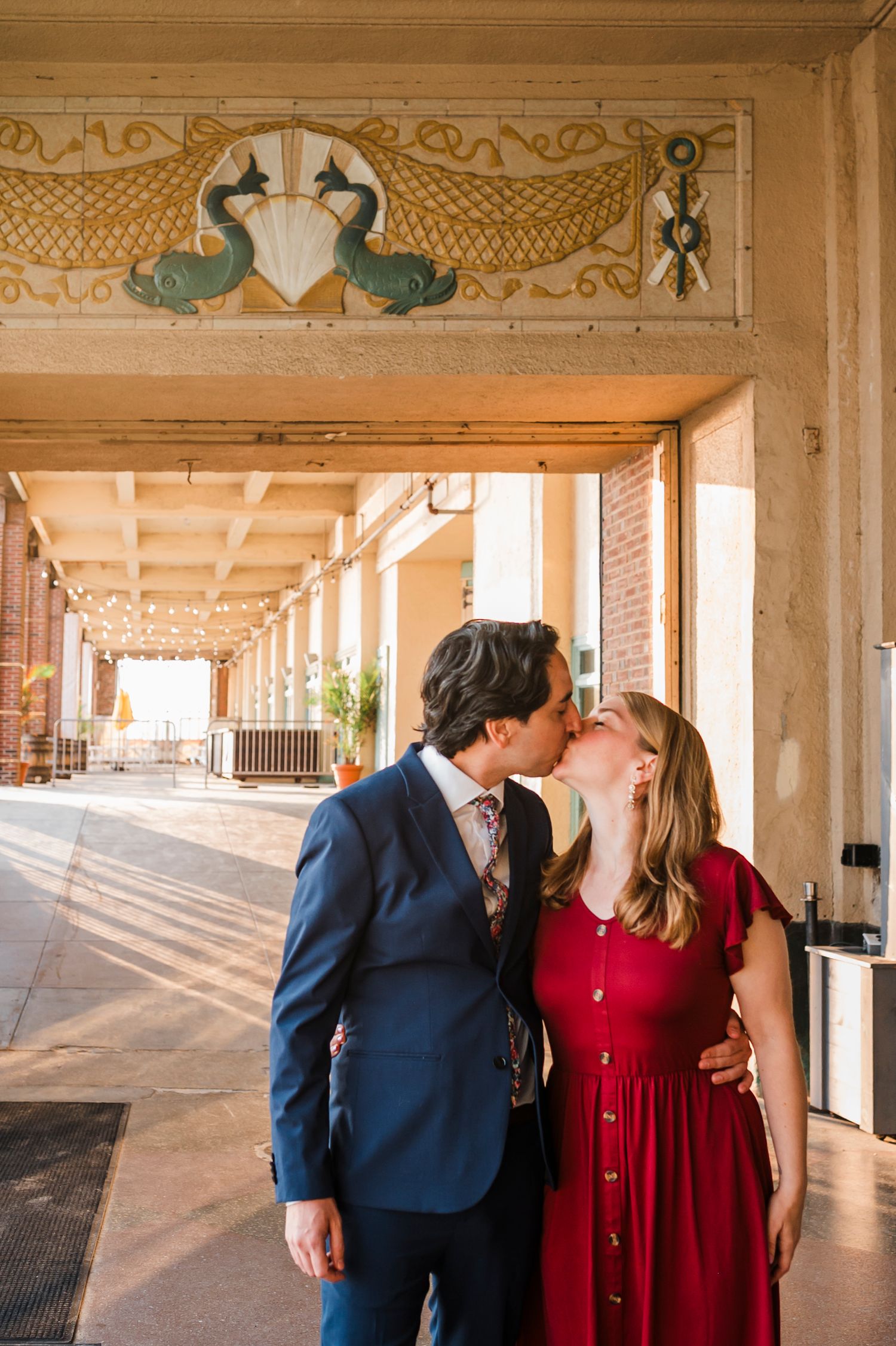 A couple embraces under an ornate colonnade with painted ceiling murals and golden details.