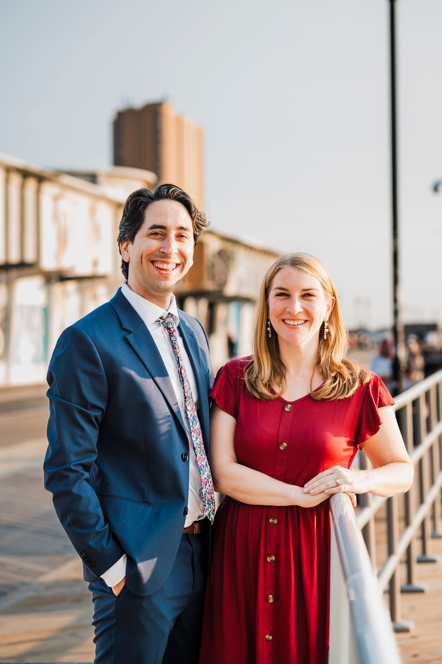 A couple poses together on a boardwalk, with the woman wearing red and the man in a navy suit.