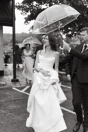 A bride in a white wedding dress walks under a clear umbrella on a rainy day during her wedding celebration.