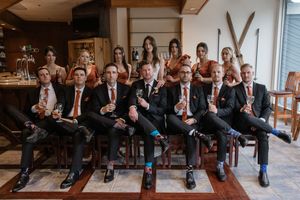 Wedding party in formal attire poses together on wooden bench with vintage skis displayed on wall behind them.
