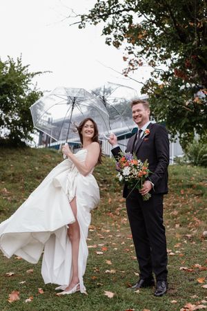 Playful wedding couple poses with umbrella in autumn setting with fallen leaves on the ground.