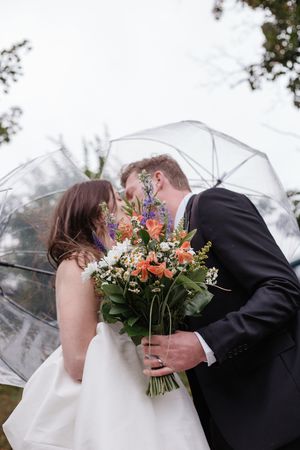 A couple shares a romantic moment under clear umbrellas while holding a vibrant wildflower bouquet.