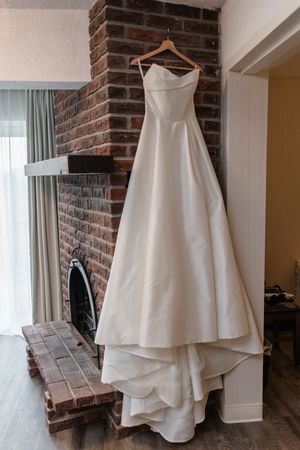 White wedding dress hanging on wooden hanger against exposed brick wall in natural lighting.