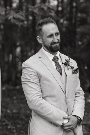 A person in a light colored suit with a boutonniere stands outdoors during a wedding ceremony.